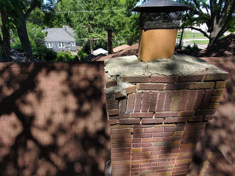 Elevated view of a brick chimney with a cracked concrete crown breaking away at the edge, displaced brick courses below and a clay flue tile with a black mesh cap