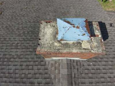 Drone view straight down onto a chimney with a badly cracked crumbling crown, missing chunks of concrete and a rusted metal cover