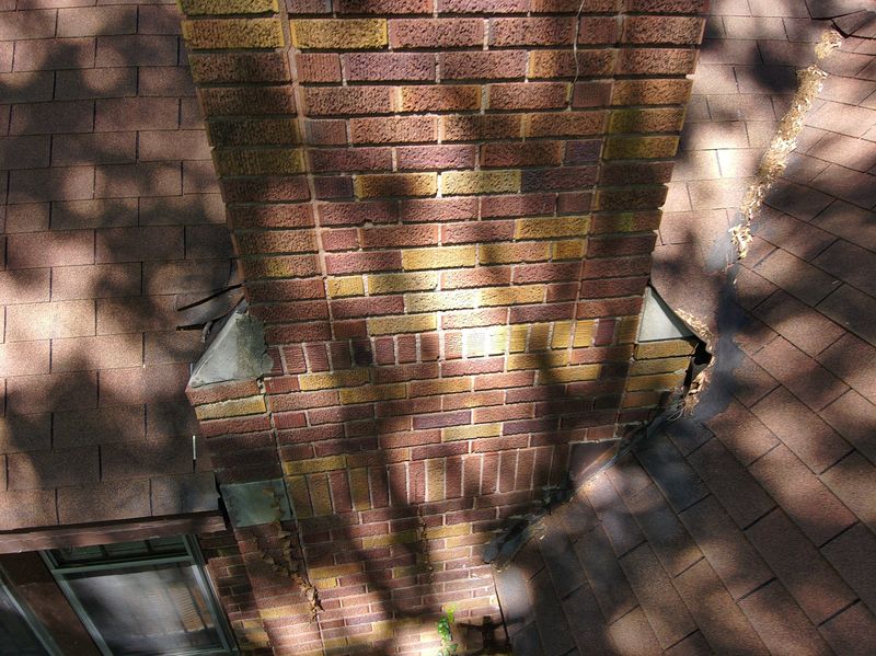 Drone view down the full face of a variegated brick chimney showing eroded mortar joints from top to bottom and aging metal at the roofline shoulders