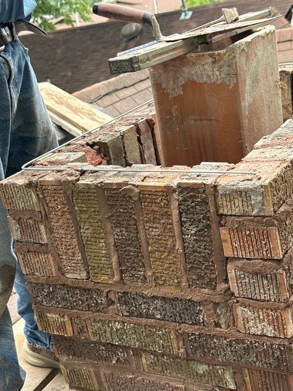 Close view of a chimney rebuild top course with steel reinforcement wire running around the perimeter beside the clay flue tile and masonry trowels resting on the brickwork