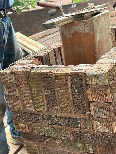 Close view of a chimney rebuild top course with steel reinforcement wire running around the perimeter beside the clay flue tile and masonry trowels resting on the brickwork
