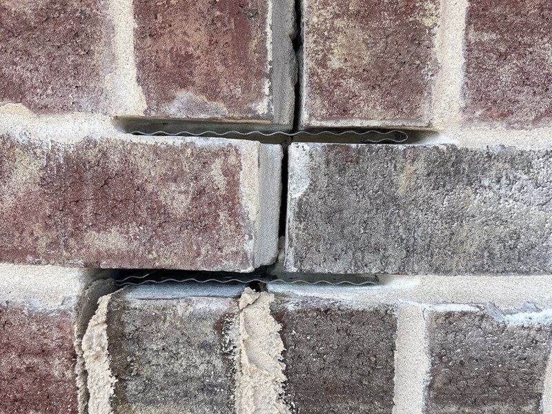 Two corrugated metal wall ties bedded in the horizontal mortar joints of red brick as the veneer is re-laid on a Madewell lintel repair