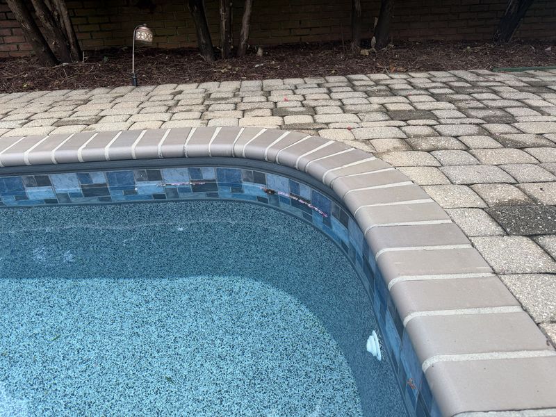 Finished brick pool coping repointed around a curved pool edge, sitting above a blue glass tile band with a paver deck behind