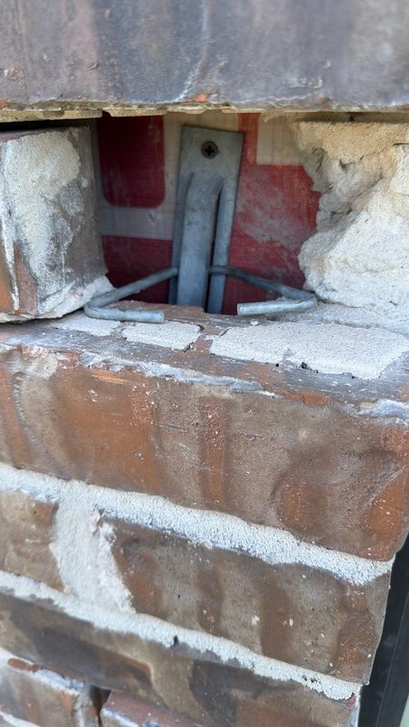 Opened brick course at a window jamb showing a metal wall tie, mortar residue, and the void behind the face brick before the opening is rebuilt
