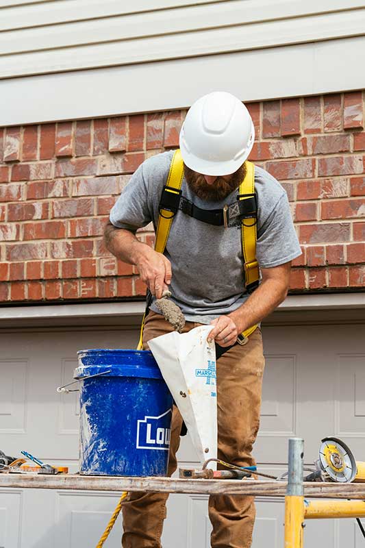 Man in white hard hat and wearing yellow harness putting mortar into a bag next to blue bucket